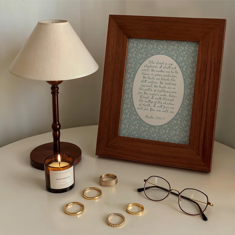 May include: A lit candle in a glass jar, a wooden framed print with text, and several gold rings are arranged on a white surface. A small lamp with a beige shade and round eyeglasses complete the scene. The framed print contains the text of Psalm 23:1-4.