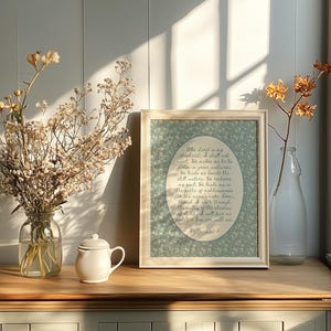 May include: A framed print with a floral background and text, a white teapot, and a clear glass vase with dried flowers sit on a wooden surface. A second vase with dried flowers is next to a window.