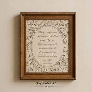 May include: A framed print with a floral pattern background and an oval-shaped central text area. The text reads "The Lord bless you and keep you...Numbers 6:24-26." The frame is a dark brown color, and the print is from "Snug Meadow Prints."