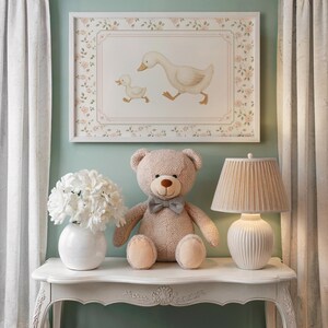 Pode incluir: Uma impress&atilde;o emoldurada de um ganso e um ganso, um urso de pel&uacute;cia com uma gravata borboleta, um vaso branco com flores e uma l&acirc;mpada com uma sombra plissada est&atilde;o em uma mesa branca. O quarto tem um esquema de cores pastel suave.