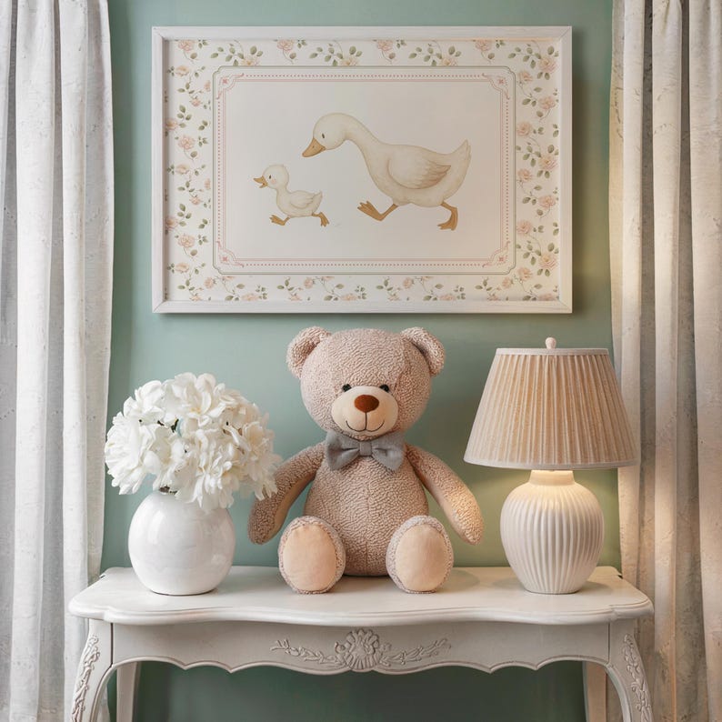 Pode incluir: Uma impress&atilde;o emoldurada de um ganso e um ganso, um urso de pel&uacute;cia com uma gravata borboleta, um vaso branco com flores e uma l&acirc;mpada com uma sombra plissada est&atilde;o em uma mesa branca. O quarto tem um esquema de cores pastel suave.