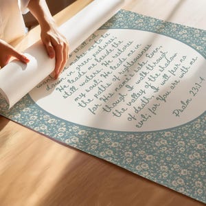 May include: A rolled-up print featuring Psalm 23:1-4 in elegant script. The print has a light blue floral border and a white background with the text. The print is being unrolled on a wooden surface.