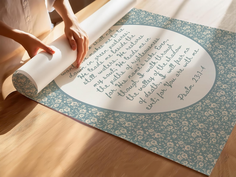 May include: A rolled-up print featuring Psalm 23:1-4 in elegant script. The print has a light blue floral border and a white background with the text. The print is being unrolled on a wooden surface.