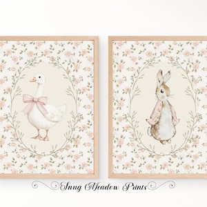May include: Two framed prints with watercolour illustrations of a goose with a pink bow and a rabbit in a pink jacket. The prints have a floral background and are framed in light wood. The text "Snug Meadow Prints" is at the bottom.