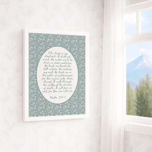 May include: A framed print featuring a floral patterned background in shades of blue and white. The print contains an oval-shaped text box with a script of Psalm 23:1-4. The frame is white, and the print is displayed near a window with a mountain view.