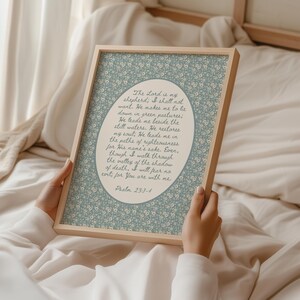 May include: A framed print featuring a floral patterned background in light blue and white. The text within the frame reads "The Lord is my shepherd...Psalm 23:1-4". The frame is made of light-colored wood.