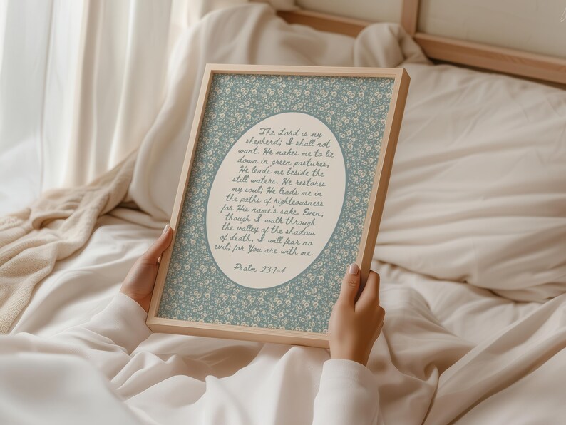 May include: A framed print featuring a floral patterned background in light blue and white. The text within the frame reads "The Lord is my shepherd...Psalm 23:1-4". The frame is made of light-colored wood.