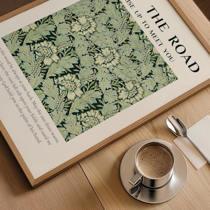 Może przedstawiać: Oprawiony wydruk z napisem "THE ROAD RISE UP TO MEET YOU" i kwiatowym wzorem w odcieniach zieleni i bieli. Filiżanka kawy na srebrnym spodku stoi na drewnianej powierzchni, z łyżeczką i małym białym kwadratem.
