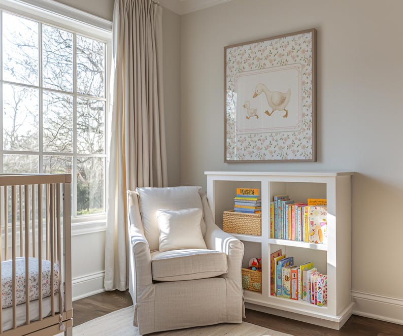 Pode incluir: Um quarto de crian&ccedil;a com um ber&ccedil;o de cor clara, uma poltrona branca e uma estante branca cheia de livros. Uma imagem emoldurada de patos est&aacute; pendurada acima da estante. O quarto tem uma grande janela com cortinas bege.