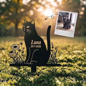 May include: A black metal cat silhouette garden stake featuring the name Luna and the years 2017-2026 engraved on the body. The design includes floral accents on both sides and is accompanied by a small photograph of a grey cat.