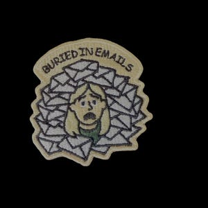 Op de afbeelding: Geborduurde patch met een cartoonfiguur begraven onder een stapel enveloppen. De patch heeft een lichtbeige rand met de woorden "BURIED IN EMAILS" bovenaan. De persoon heeft blond haar en een bezorgde uitdrukking.