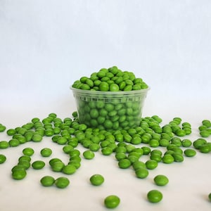 May include: A clear plastic container filled with bright green peas, with more peas scattered around it. The peas are a vibrant green color, and the container is transparent, allowing the peas to be seen clearly.