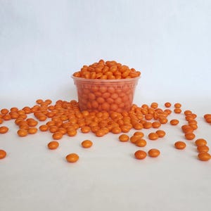 May include: A clear plastic container filled with orange candies, with more candies scattered around it. The candies are small, oval-shaped, and have a glossy finish. The background is plain white.