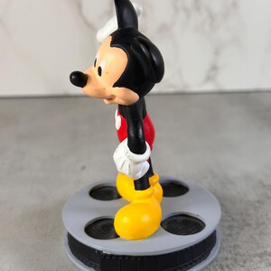 May include: A Mickey Mouse figurine standing on a circular gray base designed to resemble a film reel. The character features a black head and body, white gloves, red shorts, and large yellow shoes, captured from a side profile view.