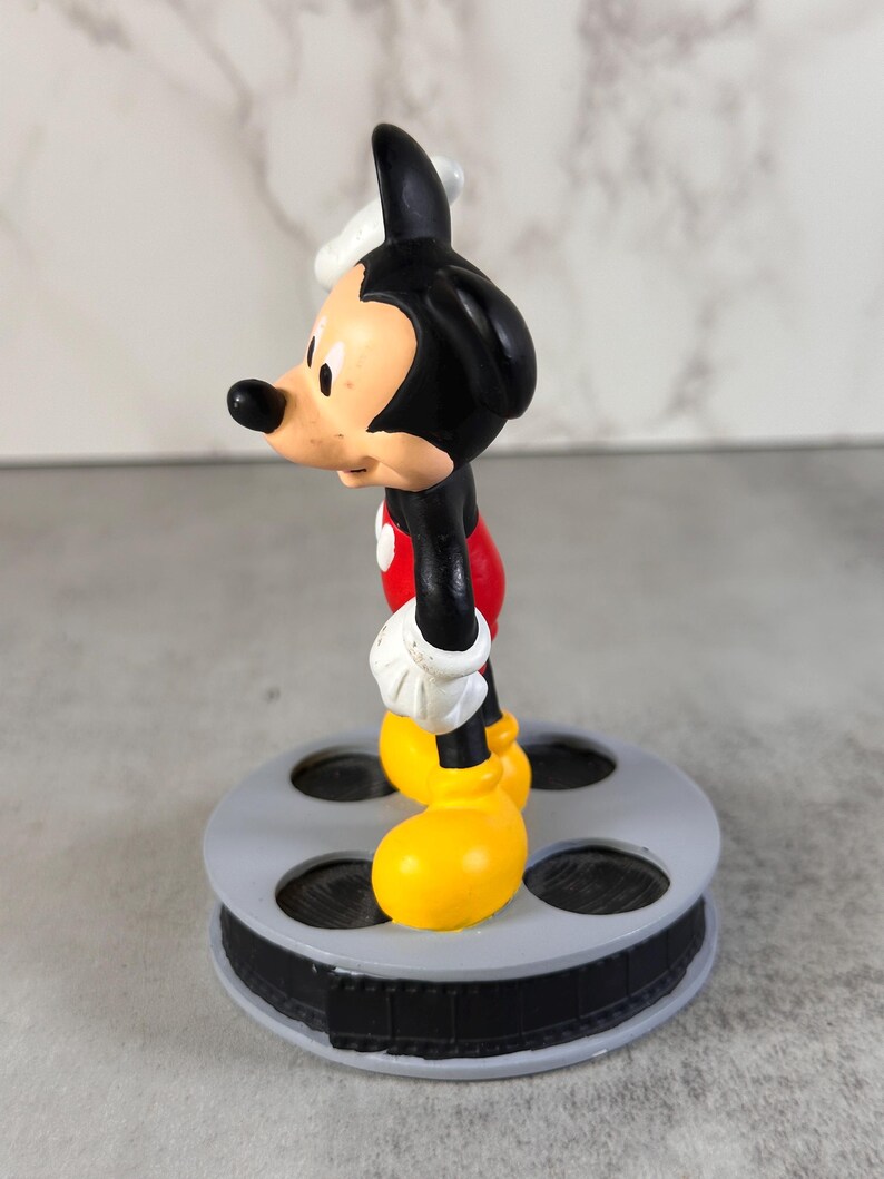 May include: A Mickey Mouse figurine standing on a circular gray base designed to resemble a film reel. The character features a black head and body, white gloves, red shorts, and large yellow shoes, captured from a side profile view.