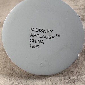 May include: A circular light gray base with black printed text that reads: &copy; DISNEY APPLAUSE TM CHINA 1999. The surface is smooth and matte, featuring a centered alignment of the manufacturer information and the production year on the bottom.