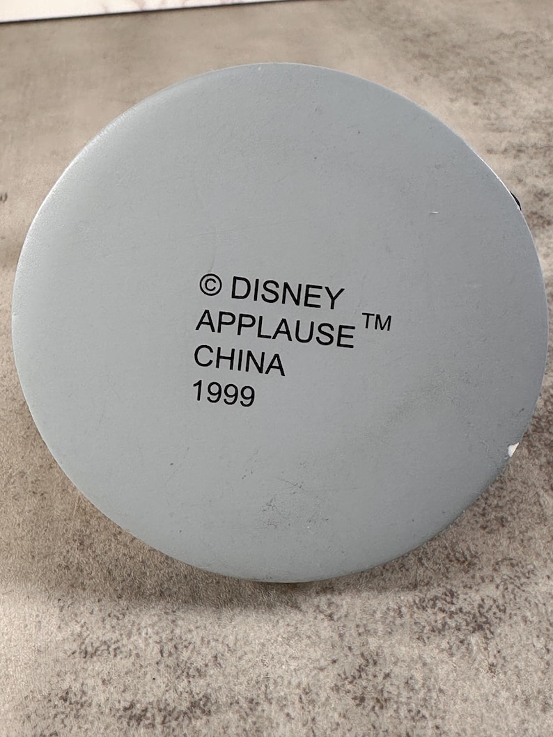 May include: A circular light gray base with black printed text that reads: &copy; DISNEY APPLAUSE TM CHINA 1999. The surface is smooth and matte, featuring a centered alignment of the manufacturer information and the production year on the bottom.