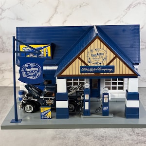 May include: A detailed model of a Ford Motor Company gas station with a blue roof and white trim. The model includes a black pickup truck with the hood open, gas pumps, and signs that read "Ford Motor Co."