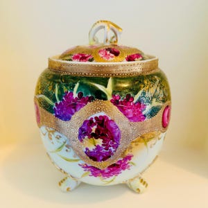 May include: A decorative porcelain jar with a lid, featuring a gold handle and trim. The jar is adorned with vibrant purple floral designs set against a green and white background. The jar rests on three small feet.