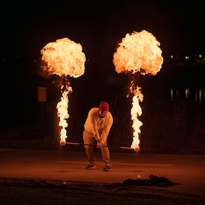 Peut inclure: Une personne en chemise jaune et chapeau rouge jongle avec le feu. Deux grandes flammes sont de chaque côté de la personne, qui tient une tige avec du feu au bout. La scène se déroule la nuit.