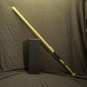 May include: A baseball bat with a yellow grip and a black and yellow patterned handle. The bat is leaning against a black rectangular box. The bat has a green ball at the end.