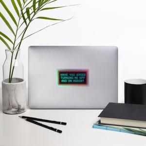 May include: A rectangular sticker with the text "HAVE YOU TRIED TURNING ME OFF AND ON AGAIN?" in green, outlined in a red and green neon border. The sticker is on a laptop.