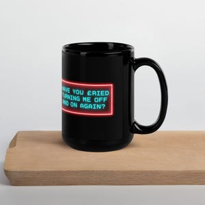 May include: Black ceramic mug with a large handle. The mug features a neon-style design with the text "HAVE YOU TRIED TURNING ME OFF AND ON AGAIN?" in a red rectangular frame. The mug is placed on a wooden surface.