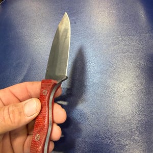 May include: A stainless steel knife with a red handle. The blade is sharp and the handle has a textured pattern with silver accents. The knife is held in a hand against a blue background.