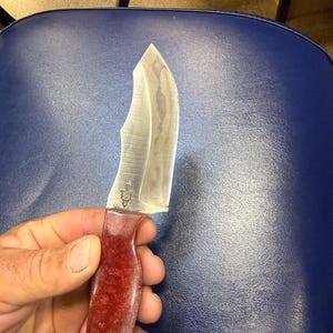May include: A handcrafted knife with a curved, silver blade and a red speckled handle. The blade has a slight curve and a pointed tip. The handle is a deep red color with a textured appearance. A small emblem is visible on the blade near the handle.