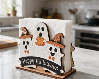 May include: A wooden napkin holder featuring three white ghost figures, two wearing orange and brown patterned hats with bows, and one holding an orange pumpkin. A black banner across the front displays the white text Happy Halloween.