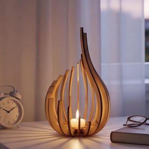May include: A light-colored wooden candle holder with a curved, slatted design that forms a teardrop shape. A small white tea light candle is lit inside the center of the structure, casting long, thin shadows across the surface it rests upon.