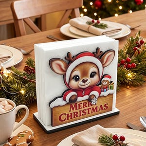 May include: A white napkin holder featuring a cartoon reindeer wearing a red hooded coat and a small teddy bear. The reindeer rests its paws on a wooden sign with the text Merry Christmas in red letters. The holder has a black base.