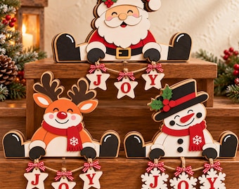 May include: Three wooden Christmas shelf sitters featuring Santa Claus, a reindeer, and a snowman. Each character has black boots and holds a string of hanging ornaments. The ornaments spell out the word JOY in red letters on star and snowflake shapes.