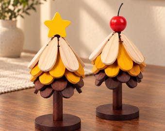 Op de afbeelding: Twee decoratieve houten boomornamenten met gelaagd, bloembladachtig gebladerte in tinten licht hout, geel en donkerbruin. De ene boom is bekroond met een gele houten ster, de andere heeft een kleine, ronde rode kers als top.