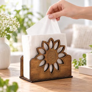 May include: A dark brown wooden napkin holder featuring a cutout sunflower design on the front panel. The holder contains a stack of folded white paper napkins, with one napkin being pulled upward from the center of the stack.