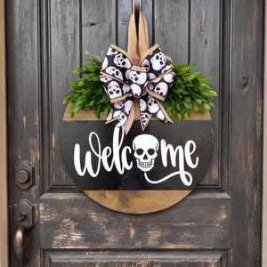 Puede incluir: Un letrero de puerta redondo de madera con una franja horizontal negra y el texto blanco "welcome", donde la letra "o" se sustituye por un gráfico de calavera blanca. Incluye un colgador de arpillera, follaje verde y un lazo con estampado de calaveras.