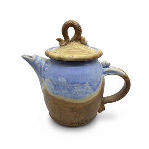 May include: Handmade ceramic teapot with a blue and brown glaze. The teapot features a curved handle and a lid with a twisted handle. The spout is angled, and the overall design is rustic and artistic.
