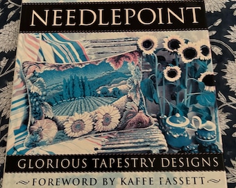May include: A hardcover book titled Jill Gordon's Needlepoint: Glorious Tapestry Designs with a foreword by Kaffe Fassett. The cover features a needlepoint pillow depicting a vineyard landscape with sunflowers, a teapot, and a teacup.