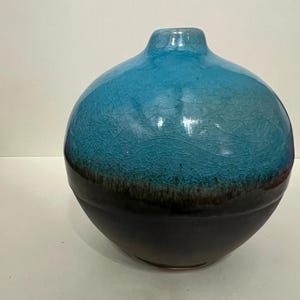 May include: A round ceramic vase with a blue and black color scheme. The top half of the vase is a vibrant blue with a cracked, textured finish. The bottom half transitions to a dark black color. The vase has a small opening at the top.