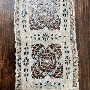 May include: A rectangular piece of off-white lace fabric featuring intricate needlework with light blue thread accents. The design includes two large circular motifs, smaller floral patterns, and a decorative scalloped border along the edges.