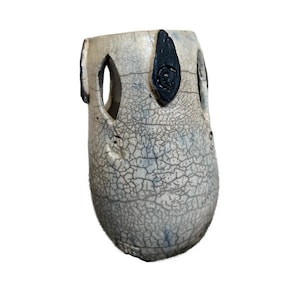 May include: A handmade ceramic vase with a crackle glaze finish. The vase is off-white with blue undertones and black accents. It features oval cutouts and decorative black shapes. The vase has a unique, textured surface.