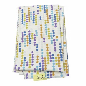 May include: Folded white fabric featuring a repeating pattern of coloured dots in blue, purple, yellow, and orange. The fabric is new. A small yellow tag reads "3yd".