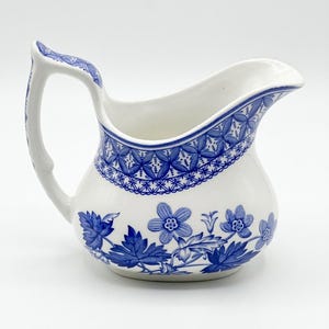 May include: White ceramic pitcher with a blue floral and geometric pattern. The pitcher has a curved handle and a spout for pouring. The blue design is on the rim, handle, and body of the pitcher.
