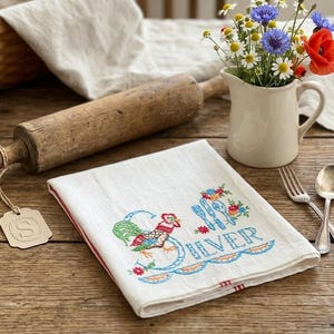 May include: A white kitchen towel featuring colorful embroidery of a rooster, floral accents, and cutlery, with the text "SILVER" stitched in blue. The towel includes a decorative tag with the letter "S" attached by a piece of twine.