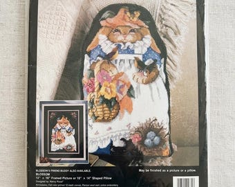 May include: A Bucilla needlepoint kit featuring a Blossom design. The kit includes a pattern for a framed picture or shaped pillow. The design features a bunny wearing a dress and holding a basket of flowers. The kit is designed by Nancy Rosa.