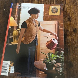 May include: A Norah Gaughan Collection Vol. 2 knitting pattern book featuring a cover image of a person wearing a textured orange button-up vest over a black shirt, holding a red speckled watering can over a potted plant, with the text 3 for all.