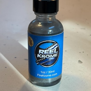 Op de afbeelding: Een heldere glazen fles met een zwarte dop en een blauw etiket. Het etiket bevat de tekst "REEL KROME" in het wit en een zilverkleurige substantie binnenin. De fles bevat 30 ml product.
