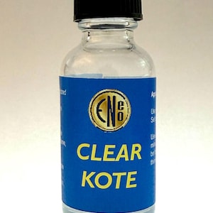 Puede incluir: Una botella de vidrio transparente con tapa negra. La botella tiene una etiqueta azul con las palabras "CLEAR KOTE" en amarillo. La etiqueta también presenta un emblema dorado. La botella está llena de líquido transparente.