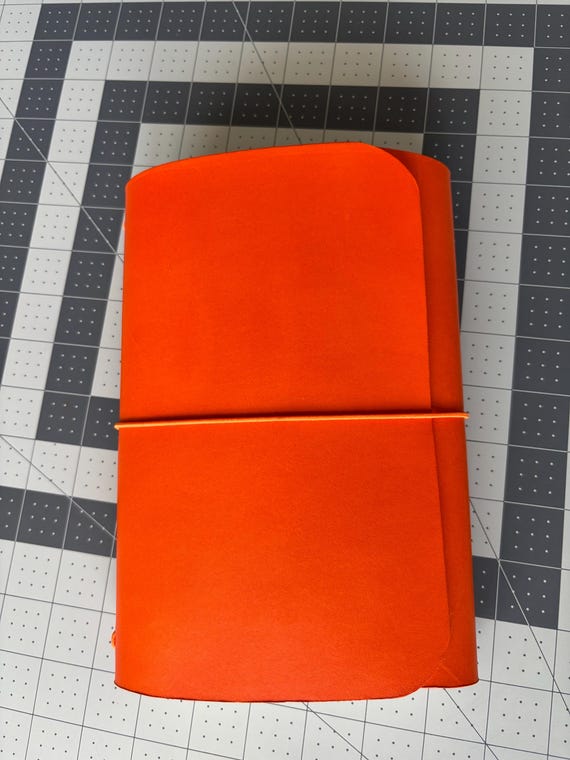 Trifold orange leather journal cover with custom cursive monogram — the structured trifold format and bold color make it stand out from the sea of plain brown leather notebooks.