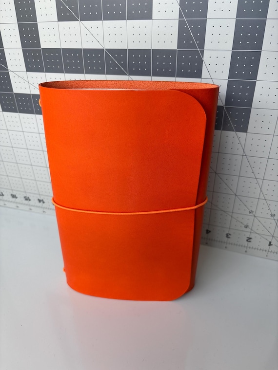 Trifold orange leather journal cover with custom cursive monogram — the structured trifold format and bold color make it stand out from the sea of plain brown leather notebooks.
